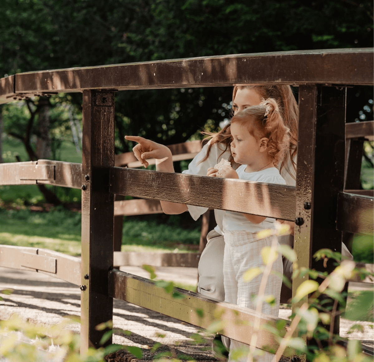 Mother and toddler standing on a wooden bridge, looking and pointing at something in the distance on a sunny day